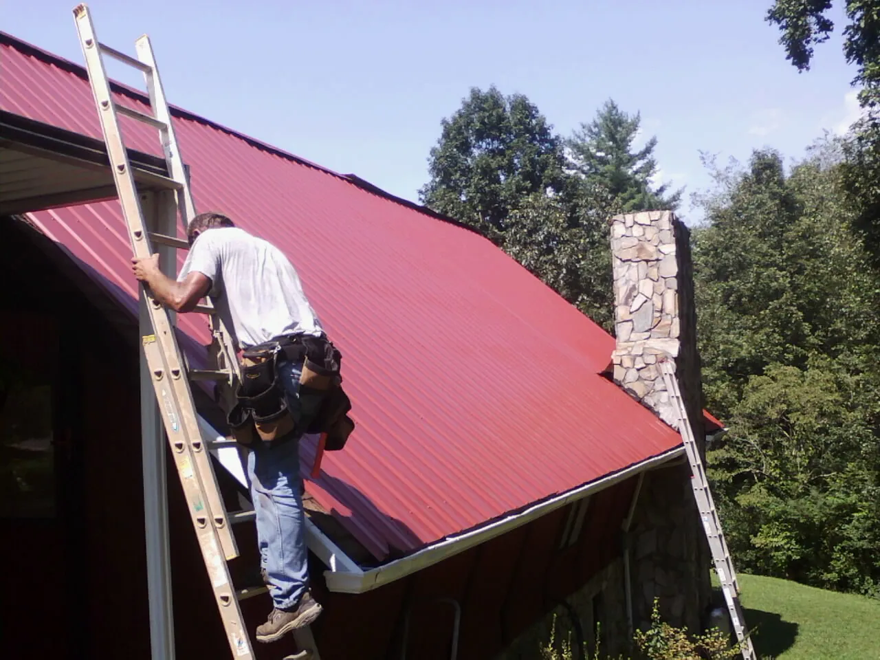 Roofer on a ladder beside a red metal roof and stone chimney — Soffit & Fascia Repair by Roofing Brothers in Reesville